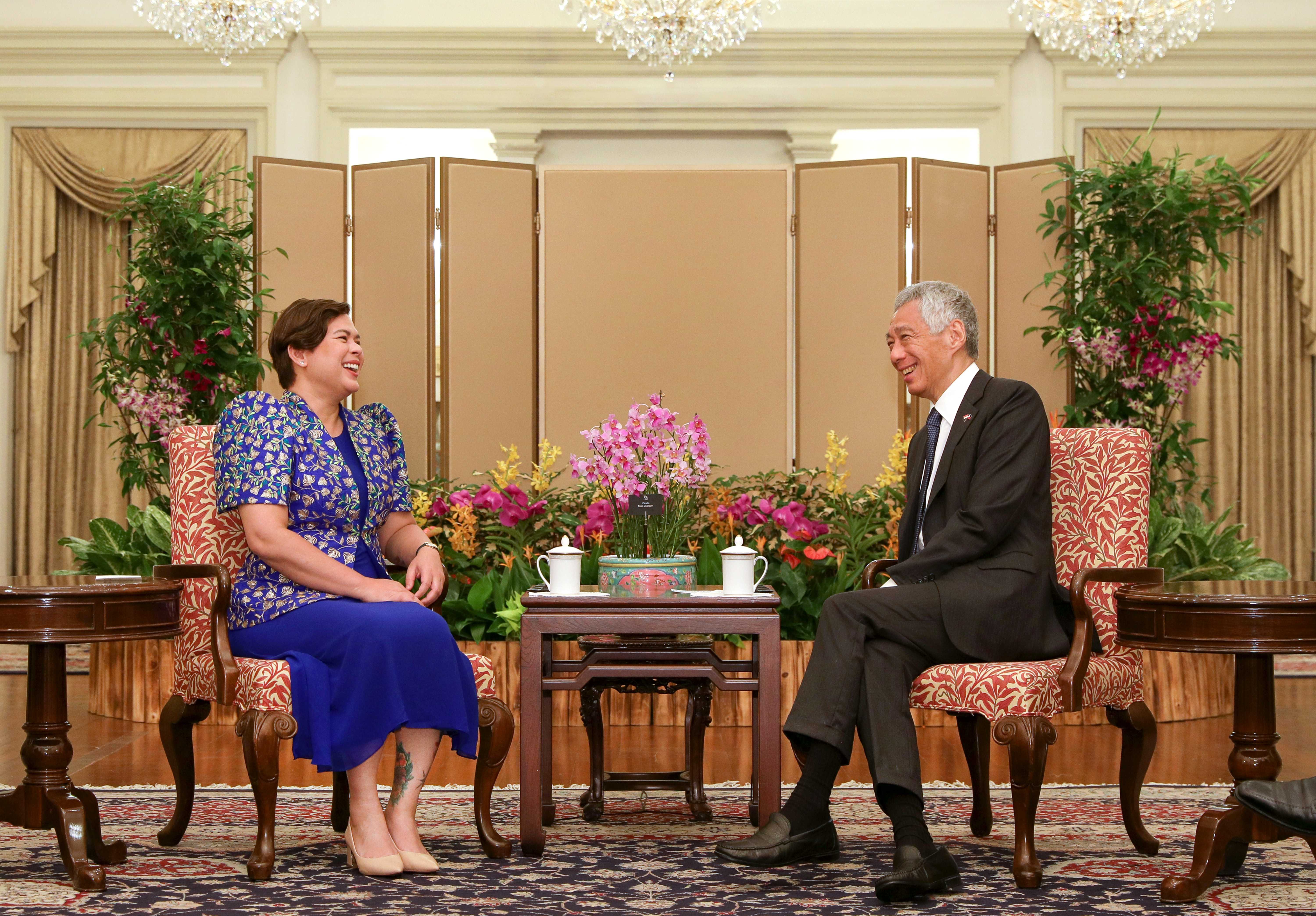 Sara Duterte and Lee Hsien Loong sit in patterned chairs, smiling at each other in ornate room.
